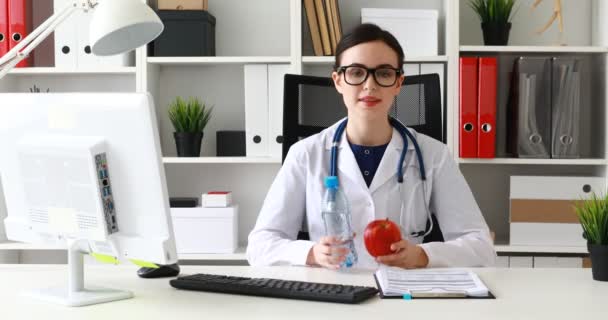 医生拿瓶水和红苹果在手 看着相机 — 图库视频影像