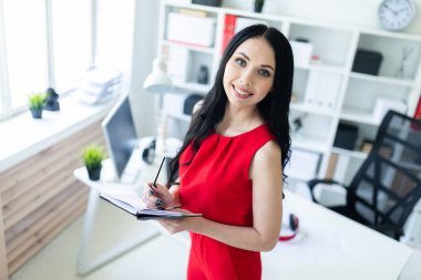 Kırmızı elbiseli güzel genç kız ofiste duran ve bir defter ve bir kalem tutan.