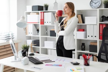 Genç bir kız ofis yakınındaki bir bilgisayar masası duruyor ve kırmızı bir fincan elinde tutan.