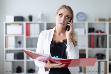 Genç bir kız ofis raf yanında duran, telefonda konuşurken ve belgeler ile bir klasör holding.