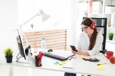 Genç bir kız kulaklık ofiste bir masada oturur ve bir telefon elinde tutan.