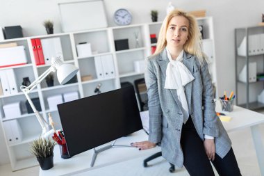 Genç kız ofiste masanın üzerine eğilerek duruyor. Yakınlarda bir monitör var.