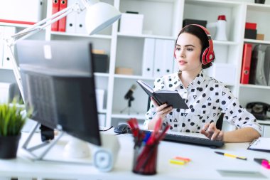 Güzel genç kız kulaklık Office masada oturur, onun elinde bir defter tutan ve klavyede yazdırır. Alan, kız odak derinliği ile fotoğraf.