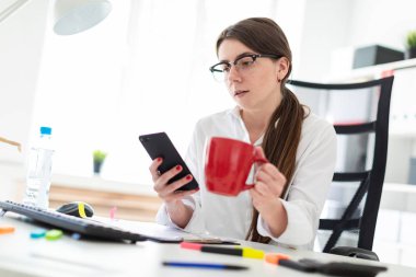 Genç bir kız bir telefon ve kırmızı bir fincan elinde tutan ofiste, bir masada oturuyor.