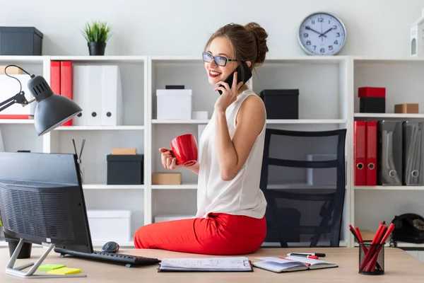 A jovem sentou-se na mesa do escritório, segurando uma xícara vermelha na mão e falando ao telefone . — Fotografia de Stock