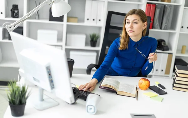 En ung pige sidder ved bordet, holder briller i hånden og arbejder ved computeren. Før pigen ligger en åben bog og et æble . - Stock-foto