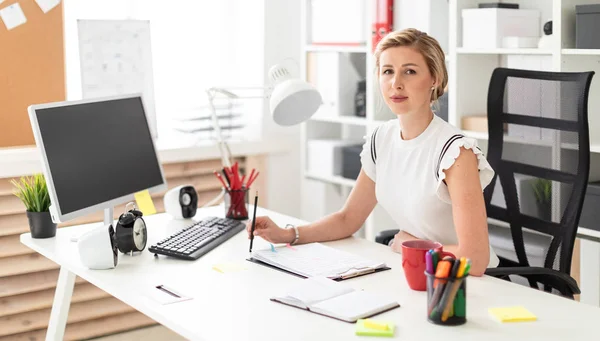 Een jong blond meisje zit achter een computerbureau in het kantoor en een potlood in haar hand houdt. — Stockfoto