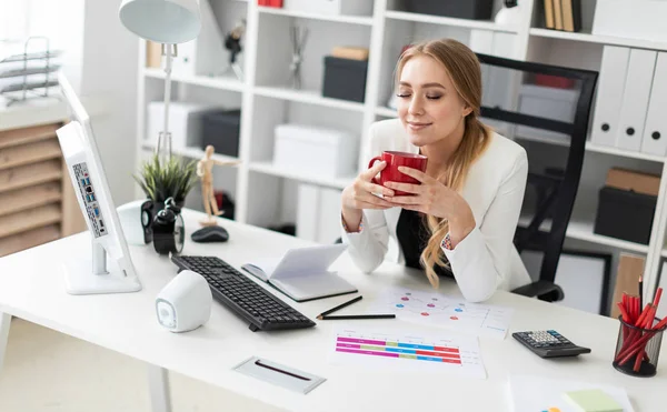 Una joven está sentada en el escritorio de la computadora en la oficina y sosteniendo una taza. Antes de que la chica en la mesa son diagramas . — Foto de Stock