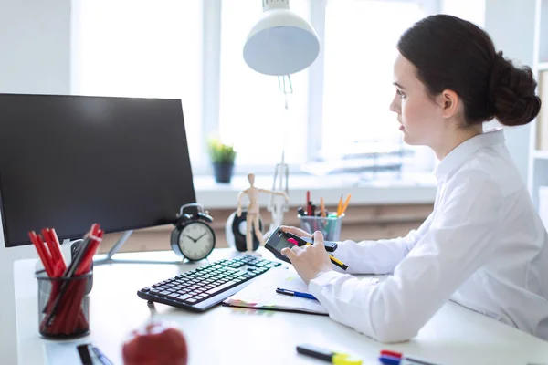 Una giovane ragazza in ufficio si siede a un tavolo, lavora con un computer, calcolatrice, documenti e tiene un pennarello e una penna nella mano sinistra . — Foto Stock