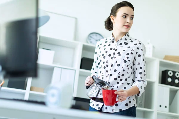 Una joven se para en la oficina cerca de la mesa y vierte el café de la cafetera en una taza roja . — Foto de Stock