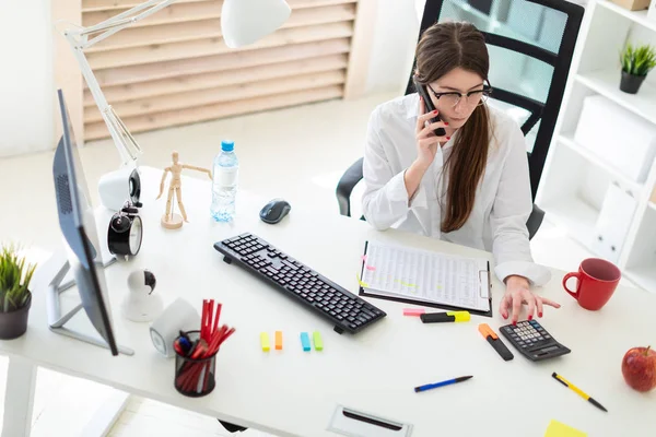 Una joven está sentada en el escritorio de la oficina, hablando por teléfono y contando con la calculadora . — Foto de Stock