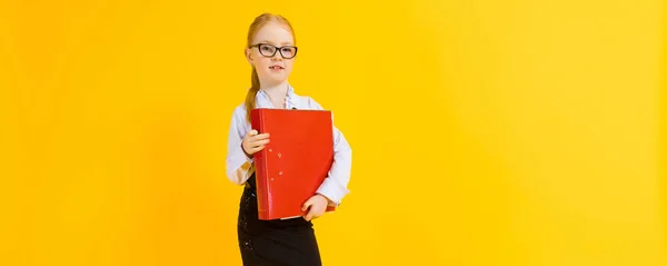 Menina com cabelo vermelho em um fundo amarelo. Uma garota encantadora em óculos transparentes está segurando uma grande pasta vermelha em suas mãos . — Fotografia de Stock