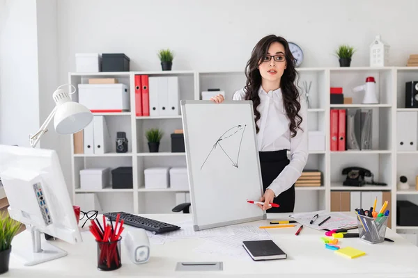 A beautiful young girl stands near an office desk and draws a magnetic marker on the magnetic board.