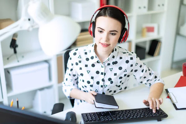 Uma linda jovem senta-se em fones de ouvido em uma mesa no escritório, segura um caderno na mão e digita o texto no teclado . — Fotografia de Stock