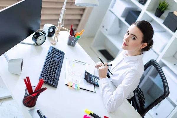 En ung flicka på kontoret sitter vid ett bord, innehar en markör och en penna i handen och arbetar med en dator, en miniräknare och dokument. — Stockfoto