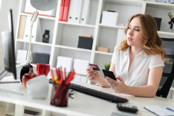 Krásná mladá dívka sedí v kanceláři, drží bankovní kartu a telefon v ruce. — Stock fotografie