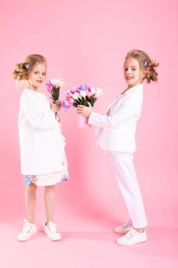 İkiz kızlar çiçek demetleri ile hafif giysiler içinde pembe bir arka plan üzerinde durmak.