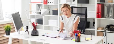 Genç sarışın kız ofiste, bilgisayar masasında oturuyor onun elinde kalem tutan ve belgeleriyle çalışma.