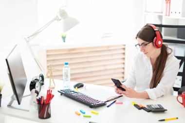 Genç bir kız kulaklık ofiste bir masada oturur ve bir telefon elinde tutan.
