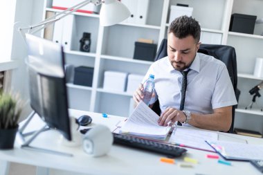 Bir adam belgeleriyle çalışma ve elinde bir şişe su tutarak ofiste, bir masada oturuyor.