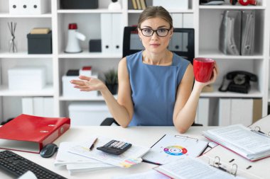 Genç bir kız ofiste bir masada oturuyor, kırmızı bir fincan elinde tutan ve belgeleriyle çalışma.