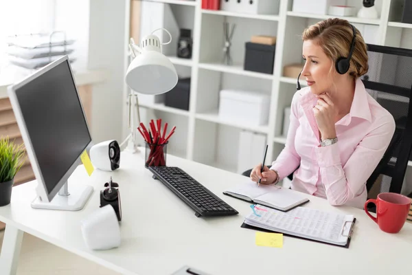 En ung flicka i hörlurarna sitter vid ett bord på kontoret, håller en penna i sin hand och arbetar med en bärbar dator och en dator. — Stockfoto