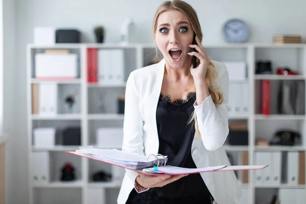 A young girl is standing in the office next to the shelving, talking on the phone and holding a folder with documents.