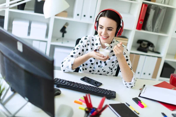 Belle jeune fille assise dans des écouteurs au bureau, mangeant du yaourt et regardant moniteur . — Photo