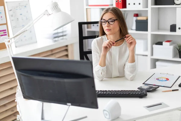 Seorang gadis muda duduk di meja di kantor, memegang pensil di tangannya dan melihat ke monitor . — Stok Foto