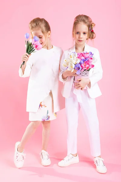 Filles jumelles en vêtements légers avec des bouquets de fleurs se tiennent sur un fond rose . — Photo