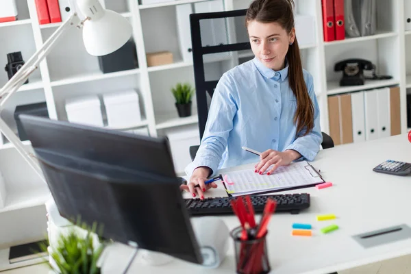 En ung flicka sitter vid ett bord på kontoret, hålla en penna i handen och arbeta vid datorn. — Stockfoto