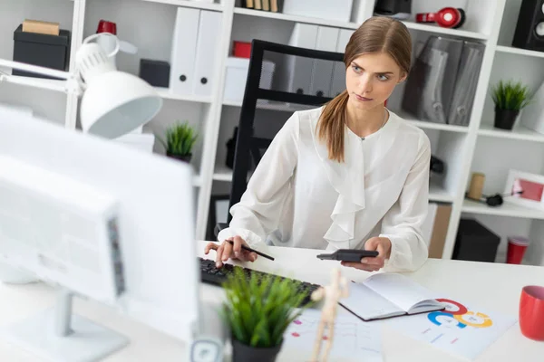 A young girl is sitting at a table in the office, holding a calculator in her hand and typing on the keyboard. — Stock Photo, Image