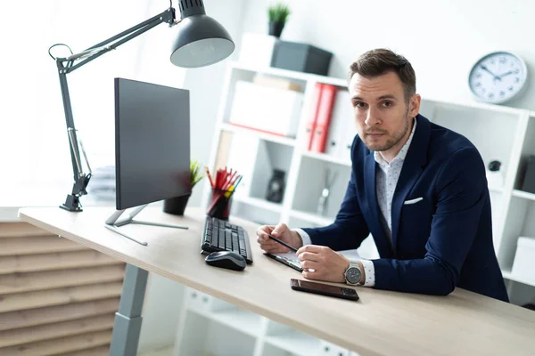 Ein junger Mann steht an einem Tisch im Büro, hält einen Bleistift in der Hand und arbeitet mit Dokumenten und einem Computer. — Stockfoto