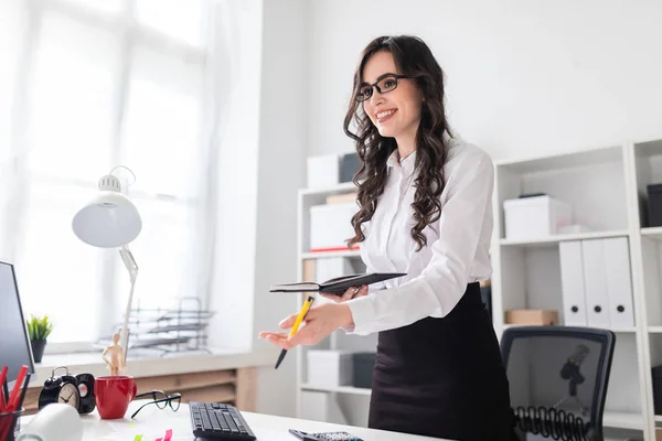 Een mooi jong meisje staat in de buurt van een bureau en houdt een pen en een laptop in haar handen. Het meisje onderhandelt. — Stockfoto