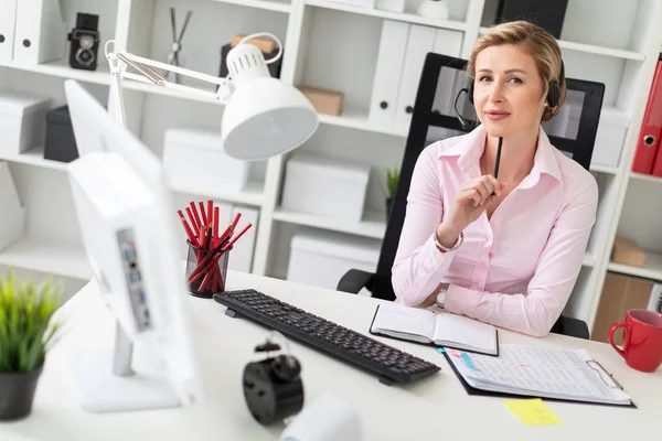 Een jong meisje in de koptelefoon zit aan een tafel in het kantoor, houdt een potlood in haar hand en werkt met een notebook en een computer. — Stockfoto