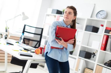 Ofiste genç bir kız, bir masaya eğilmiş duruyor ve bir telefon ve belgeler ile bir klasör tutuyor.