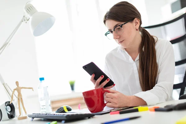 En ung flicka sitter vid ett bord på kontoret, hålla en telefon och en röd kopp i handen. — Stockfoto