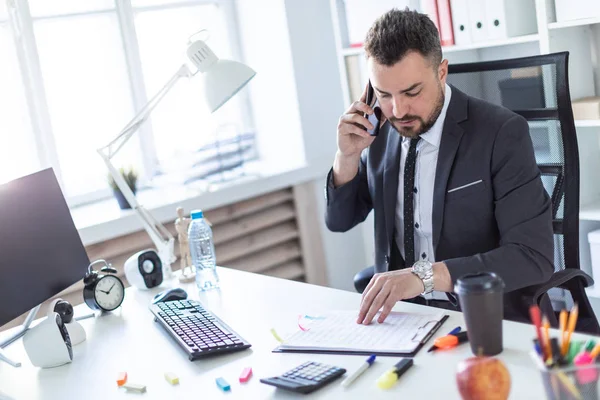 stock image A man is sitting in the office at the table, talking on the phone and flipping through documents.