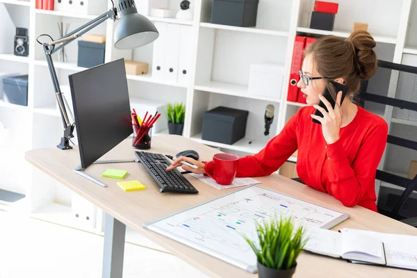 Een jong meisje is zitten op het Bureau in het kantoor, een marker holding in haar hand, praten over de telefoon en typen op het toetsenbord. Voor het meisje liggen een notebook en een magneetbord. — Stockfoto