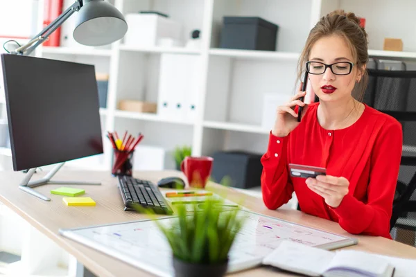 Una joven está sentada en el escritorio de la oficina, con una tarjeta bancaria y un teléfono en la mano. . — Foto de Stock