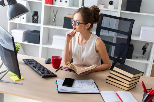 En ung flicka sitter vid en dator skrivbord och håller en öppen bok i händerna och ser på bildskärmen. — Stockfoto