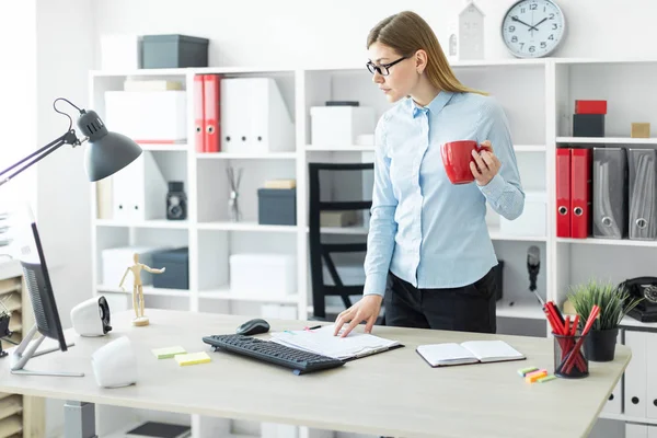 Una joven en gafas está de pie cerca de la mesa, sosteniendo una taza roja en su mano y escribiendo en el teclado . — Foto de Stock