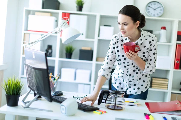 En ung jente står på kontoret ved bordet med et rødt krus i hånden og skriver på tastaturet. . – stockfoto