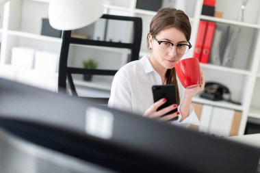 Genç bir kız bir telefon ve kırmızı bir fincan elinde tutan ofiste, bir masada oturuyor.