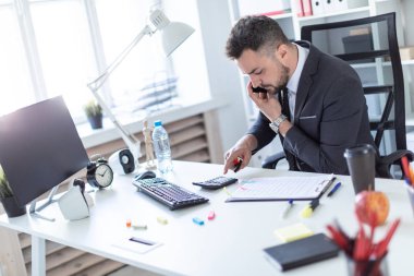 Bir adam ofiste masa başında oturan, telefonda konuşurken, elinde kalem tutan ve hesap makinesinde sayma.