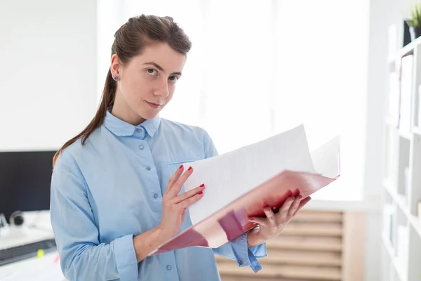 En ung flicka på kontoret står nära vindskyddet och bläddrar igenom mappen med dokument. — Stockfoto