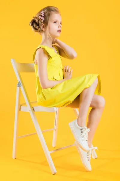 Ein Mädchen sitzt auf einem weißen Stuhl auf gelbem Hintergrund. — Stockfoto