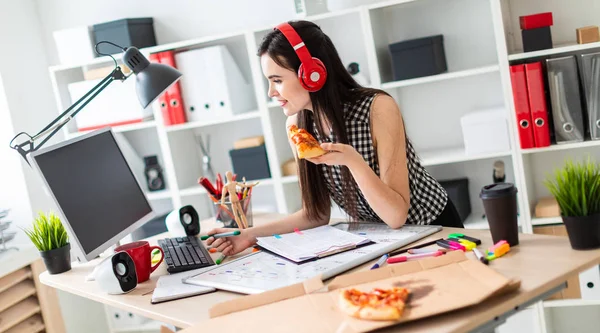 Uma jovem está parada perto de uma mesa, segurando um marcador verde e um pedaço de pizza na mão. Antes que a rapariga em cima da mesa seja um quadro magnético. Na cabeça da menina usando fones de ouvido . — Fotografia de Stock