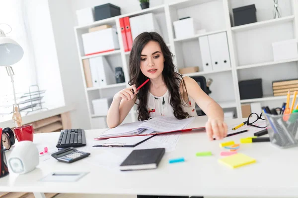 Een mooi jong meisje zit bij een bureau houdt een pen in de buurt van haar gezicht en bereiken voor heldere stickers. — Stockfoto