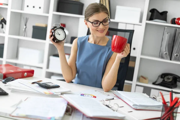 Una joven está sentada en una mesa en su oficina, sosteniendo un despertador y una taza roja . — Foto de Stock